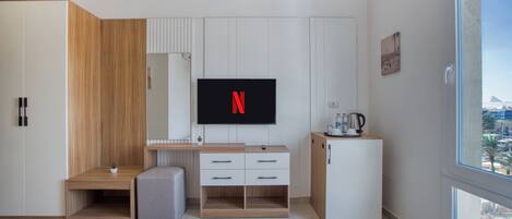 A modern hotel room with a wooden desk, a flat-screen TV, a minibar, and a large window offering a city view.