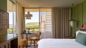 Ett hotellrum med en stor säng, ett skrivbord, en stol, ett litet bord med en vas och utsikt över en stadspanorama genom stora fönster.