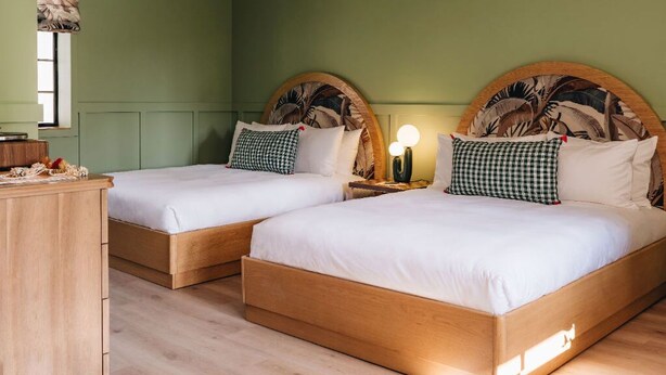 Ein Zimmer mit zwei Holzbetten, beide mit einem Kopfstück mit Palmenblattmuster. Ein Holzschrank befindet sich links und ein Nachttisch mit Lampe zwischen den Betten.