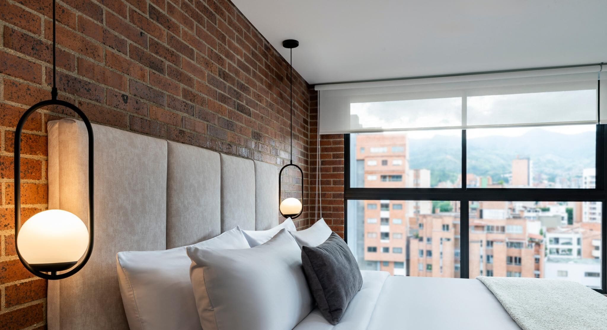 A modern bedroom with a brick wall, a large window with a city view, and a bed with multiple pillows.