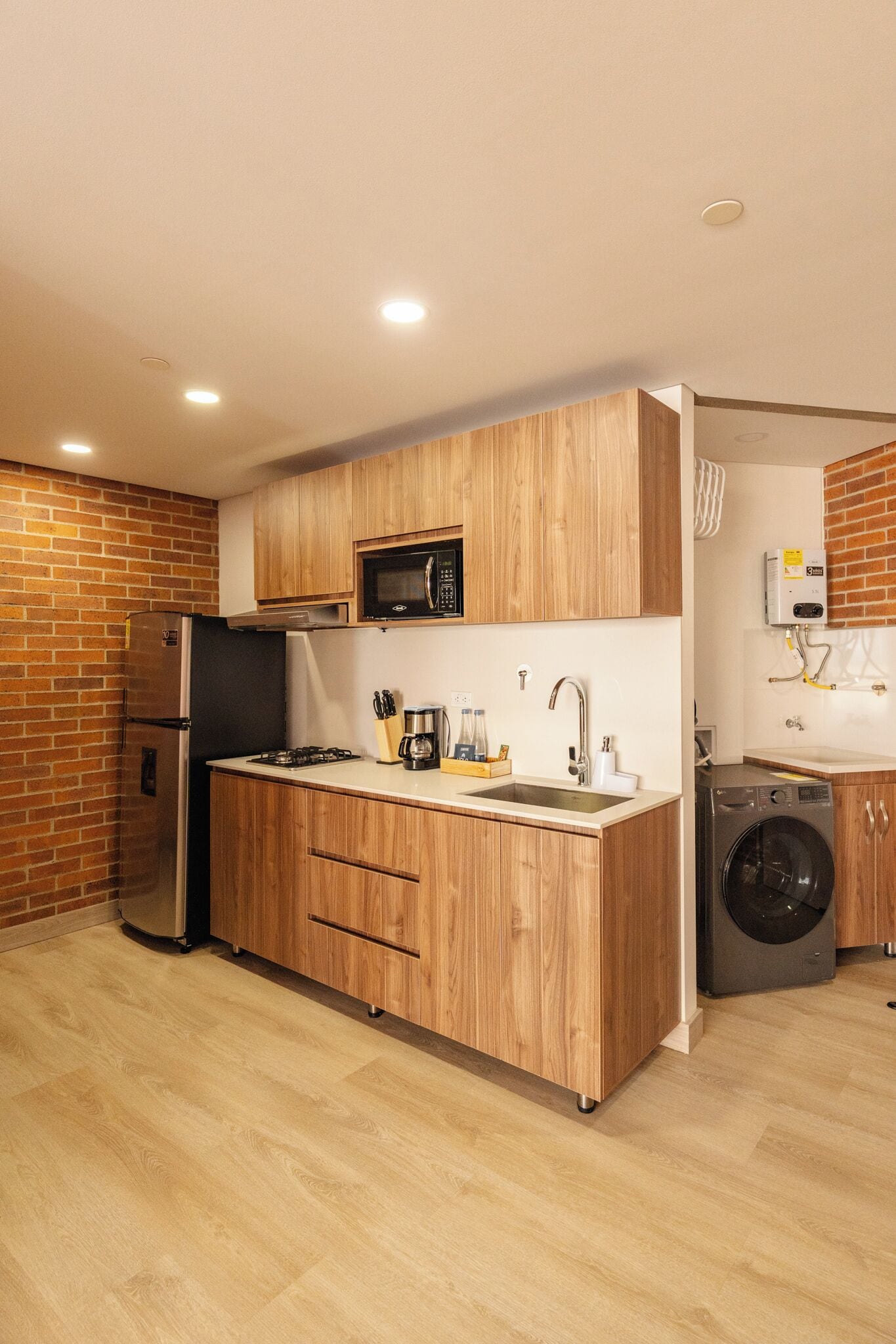 A compact kitchen with wooden cabinets, a built-in microwave, a stainless steel refrigerator, a sink, and a washing machine.