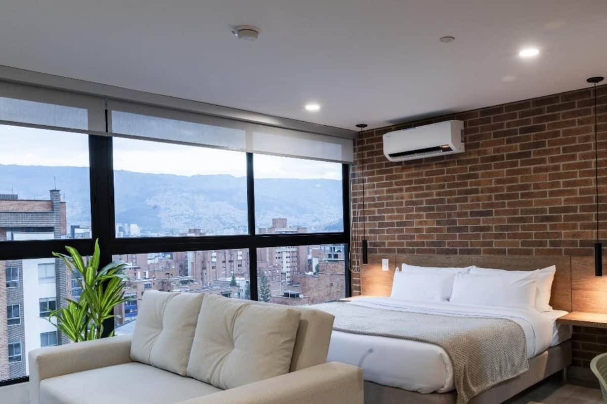 A modern hotel room with a large window, a bed, a sofa, and a brick wall.