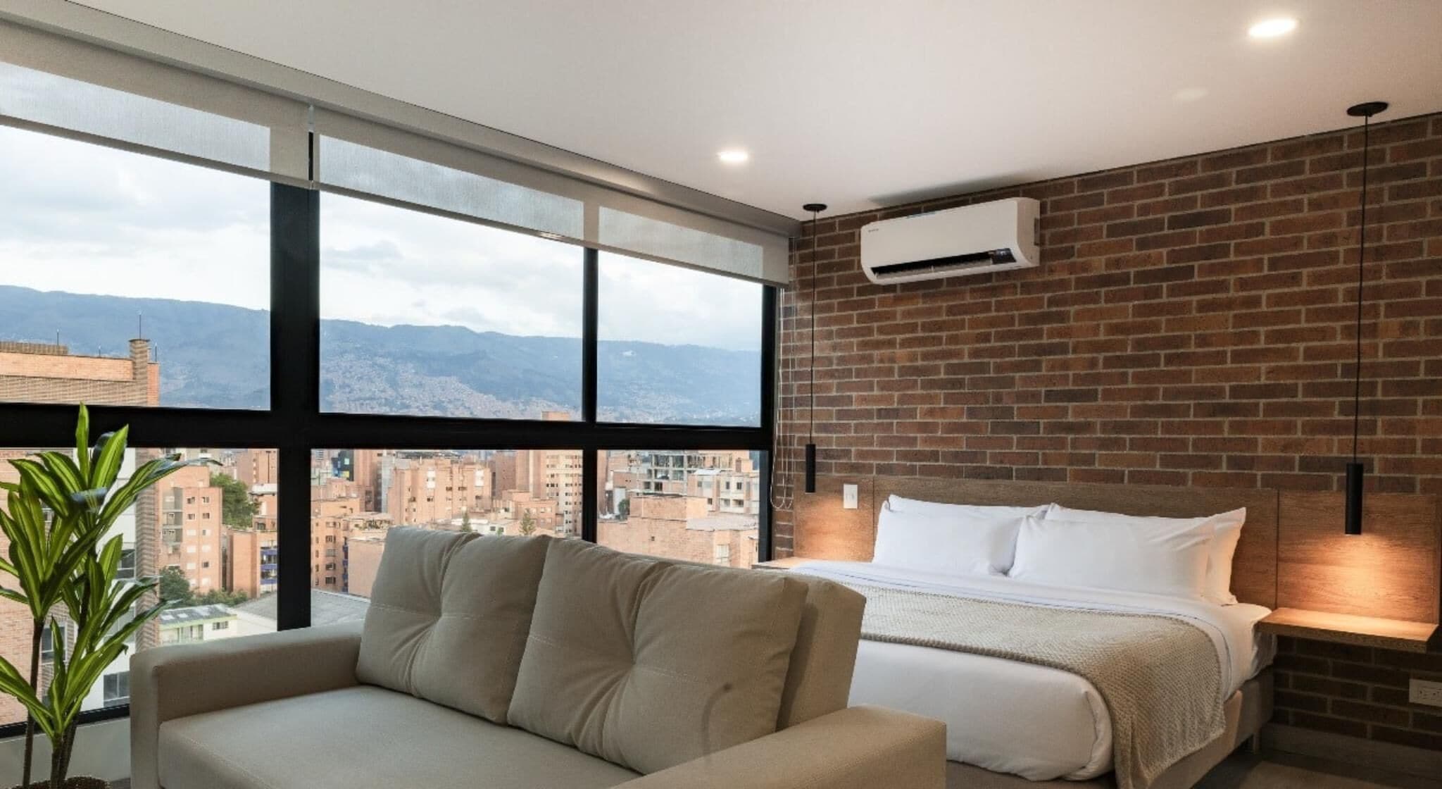Une chambre d’hôtel moderne dotée d’une grande fenêtre donnant sur la ville, d’un lit avec du linge de maison blanc, d’un canapé beige et d’un mur décoratif en briques.
