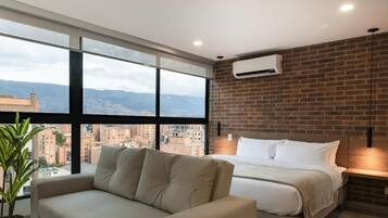 A modern hotel room with a large window offering a city view, a bed with white linens, a beige sofa, and a brick accent wall.