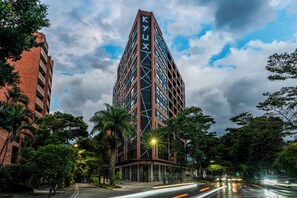 A tall, modern building with the name 'KYUX' on its facade, surrounded by lush greenery and a wet street reflecting city lights. - Kyux by Jalo (Medellín)