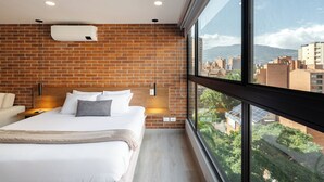 A modern hotel room with a brick wall, a large bed, a nightstand, and a view of the city through large windows. - Kyux by Jalo (Medellín)