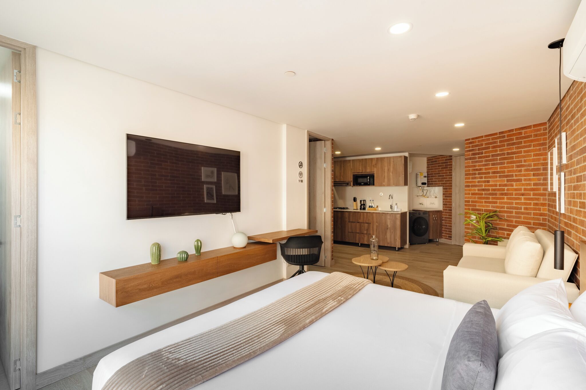 A modern hotel room with a large bed, a wooden shelf with decorative items, a black chair, a small round table, and a kitchenette in the background.