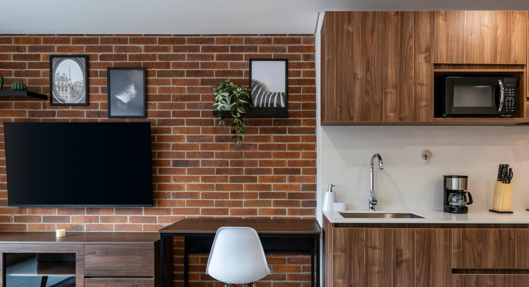 A modern kitchen with a brick wall, wooden cabinets, a flat-screen TV, a sink, and a coffee machine.