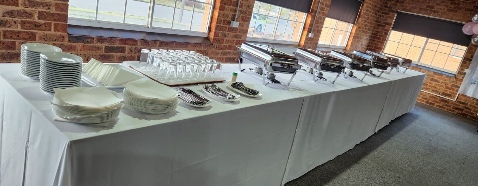 A buffet setup with plates, glasses, cutlery, and hot food stations.