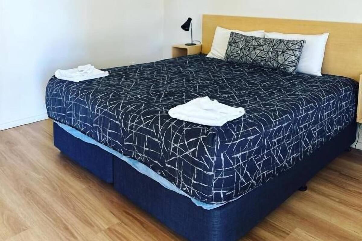 En snyggt bäddad säng med ett geometriskt mönstrat överkast, en enda kudde och en sänglampa.