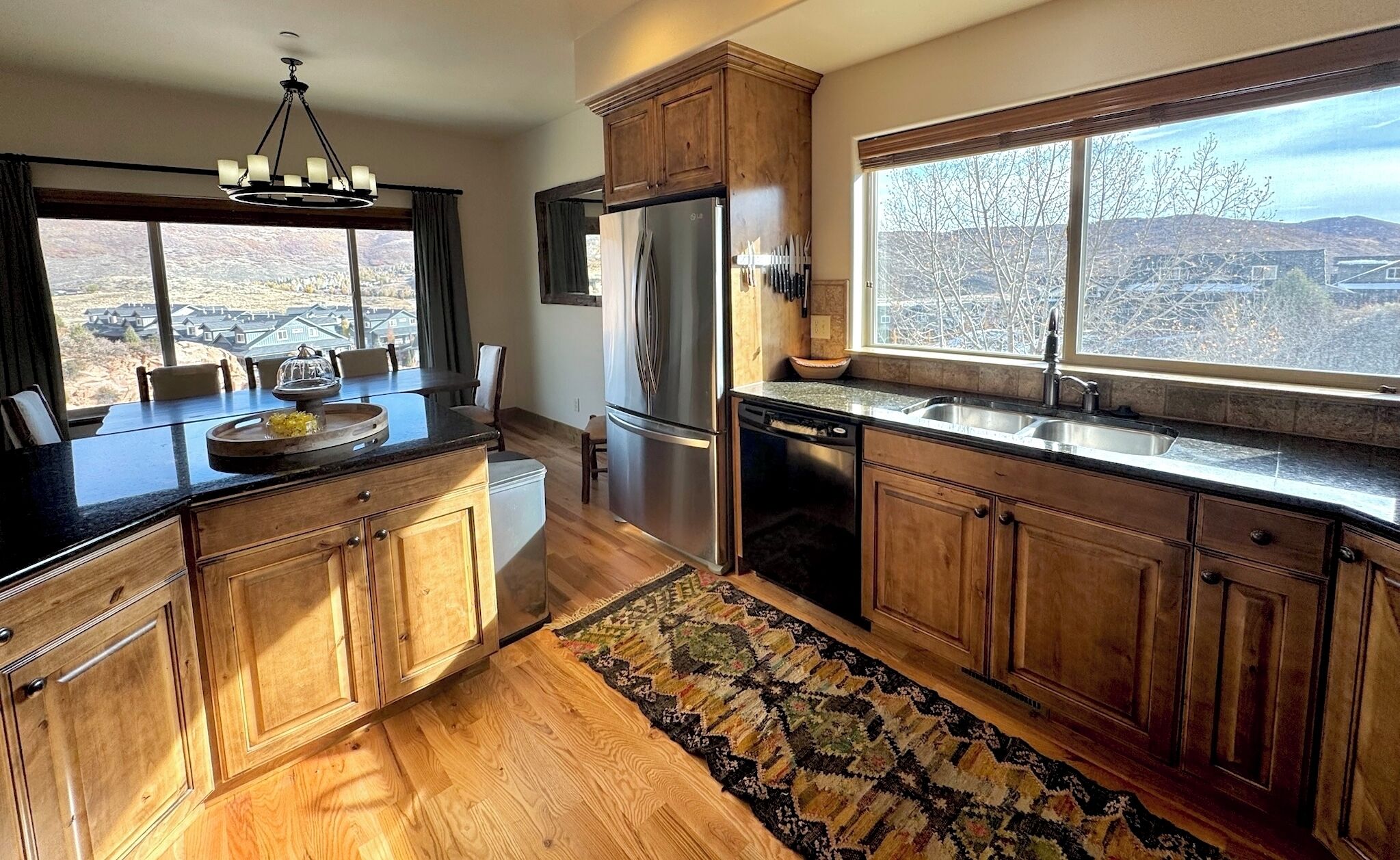 A kitchen with wooden cabinets, a stainless steel refrigerator, a dishwasher, and a window with a view of houses and trees.