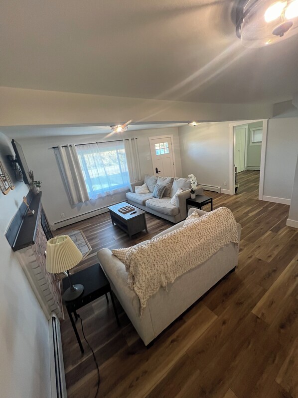 A spacious living room with a large sofa, a coffee table, and a lamp. There is a door leading to another room and a window with curtains.