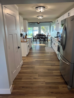 A modern kitchen with wooden flooring, white cabinets, a stainless steel refrigerator, and a dining area with a view of the outdoors.
