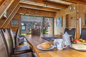 Un intérieur en bois avec une table à manger, des chaises, une cafetière et une assiette contenant du pain et des fruits.