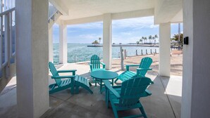 A covered outdoor seating area with turquoise Adirondack chairs and a small round table, overlooking a beach with palm trees and a pier. - Post Card Point #319 - Ocean Views! (Cudjoe Key)