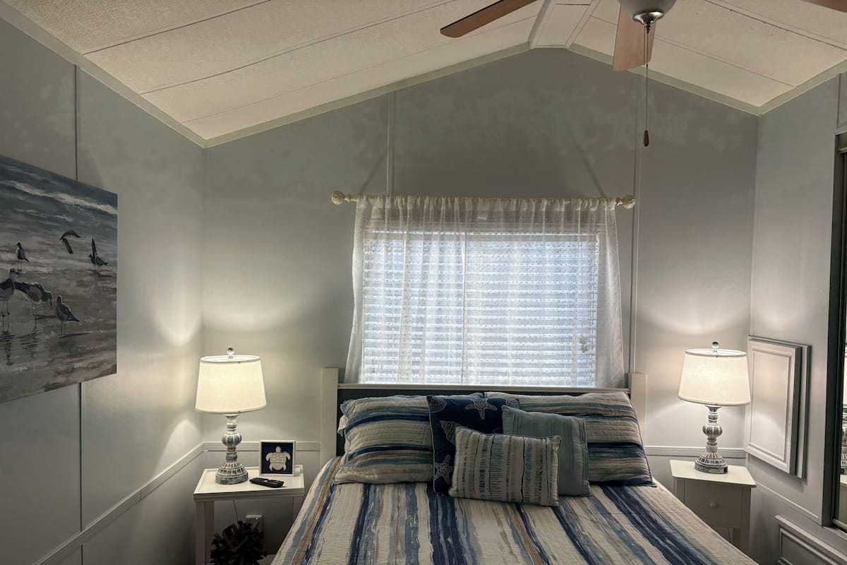 A bedroom with a bed, two bedside tables with lamps, a window with sheer curtains, and a ceiling fan.