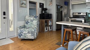 Een moderne keuken met witte kasten, een ingebouwde magnetron, een roestvrijstalen koelkast en een gasfornuis. Er staat een houten barstoel en een blauwe fauteuil.