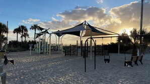 Een strand met een speeltuin, inclusief schommels en klimconstructies, onder een grote parasol.
