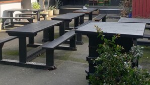 Picknicktafels buiten opgesteld in rijen, met potplanten en een muur waarop een bord hangt.
