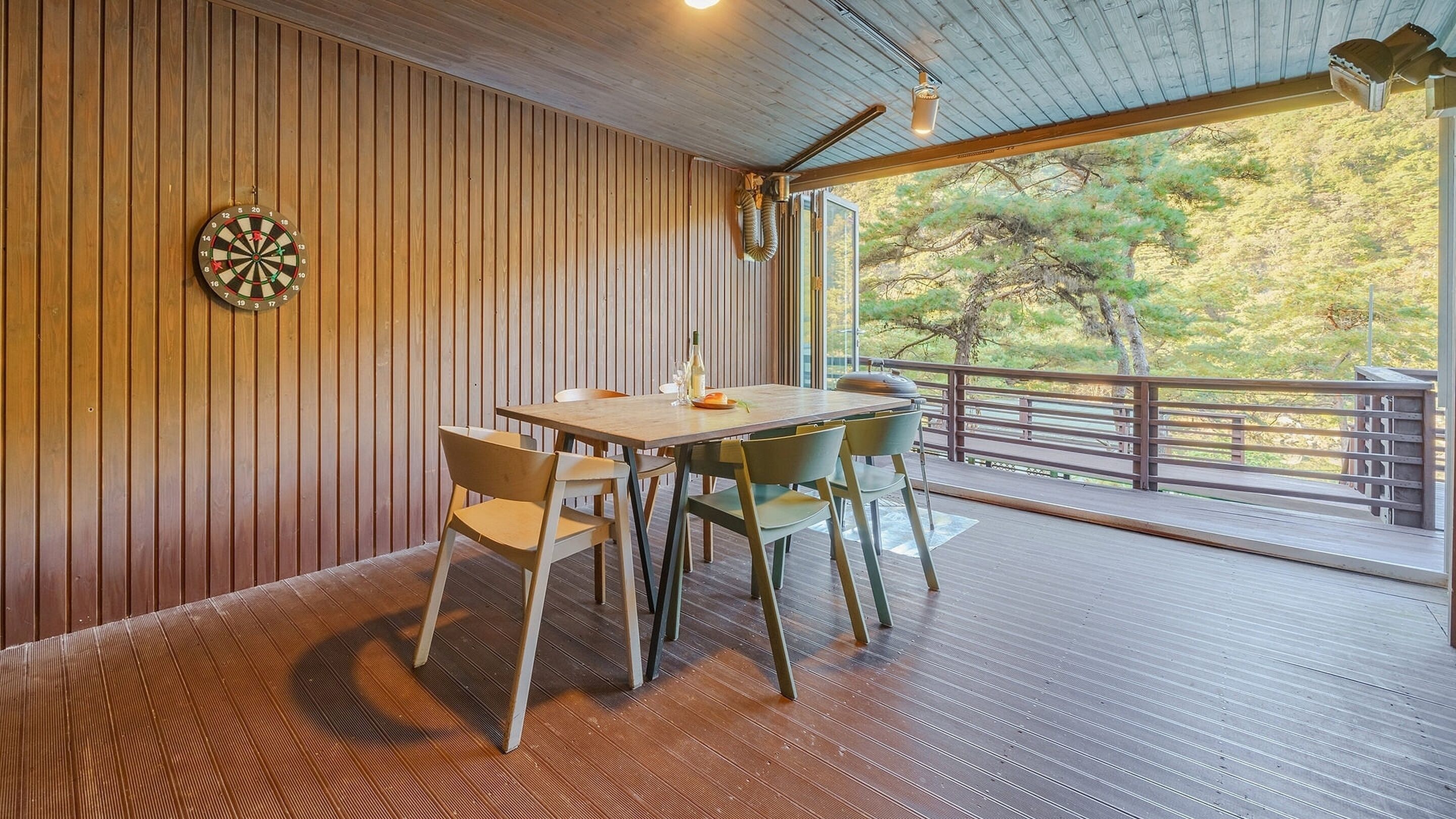 A dining area with a wooden table and chairs, a dartboard, and a view of trees through large windows.
