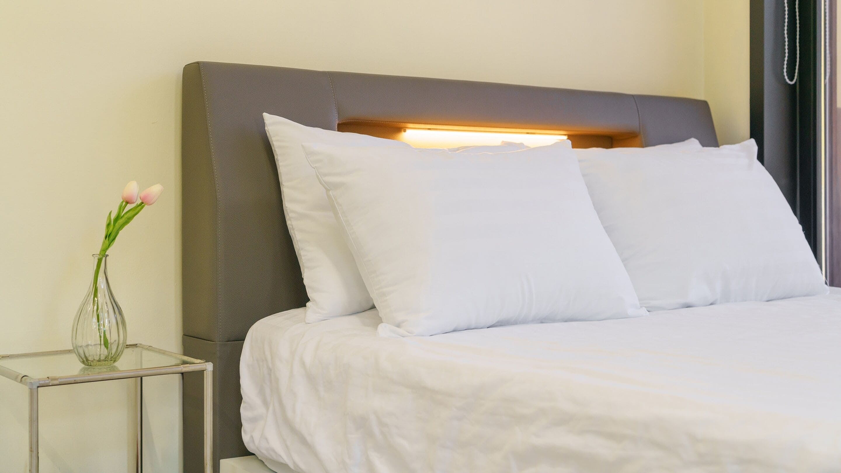 Una cama bien hecha con sábanas y almohadas blancas, un jarrón de cristal transparente con flores sobre una mesita de noche, y un cabecero moderno con iluminación integrada.