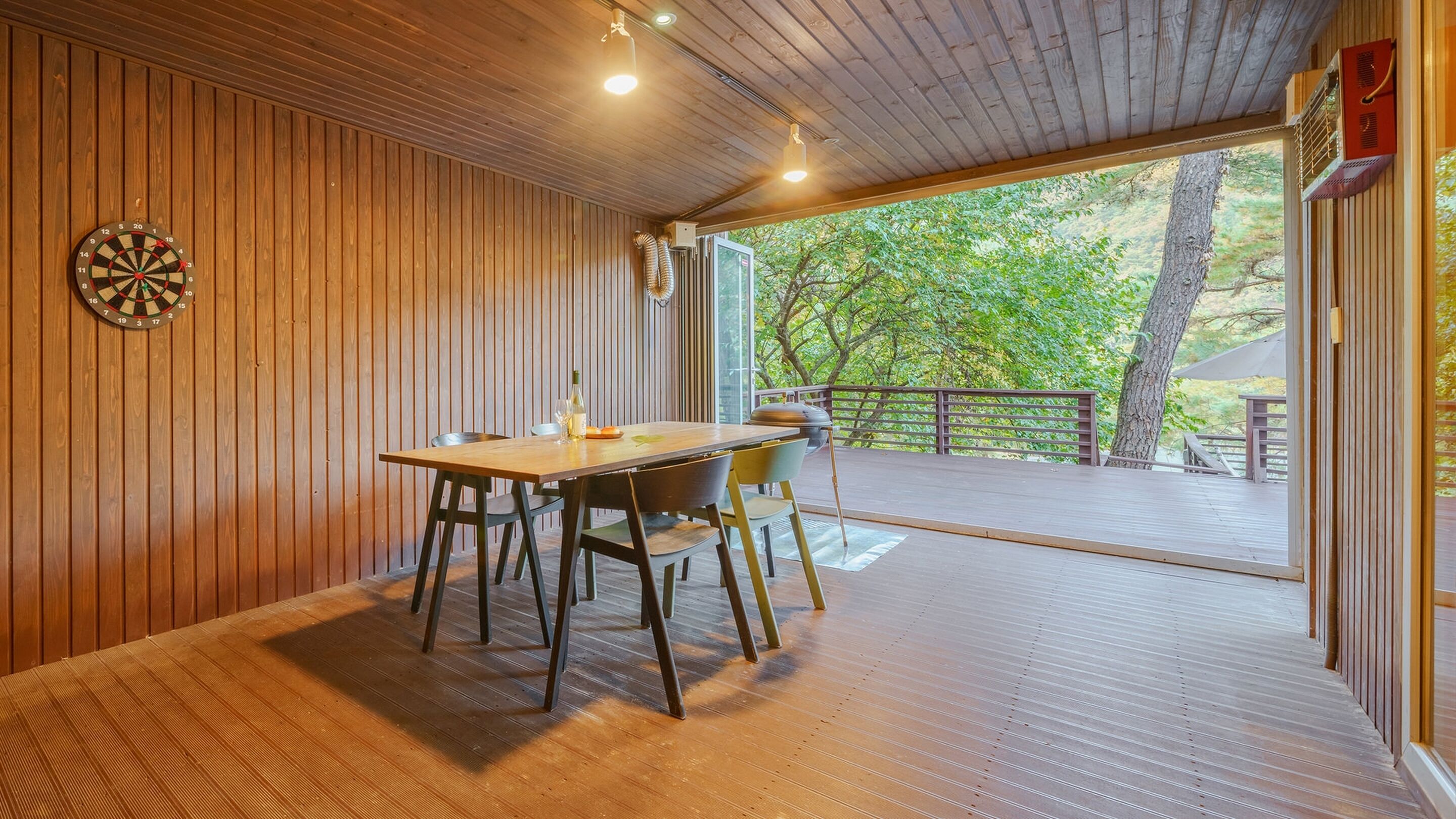 Un interior de madera con una mesa y sillas, un dardo y un ventanal con vistas a una terraza.