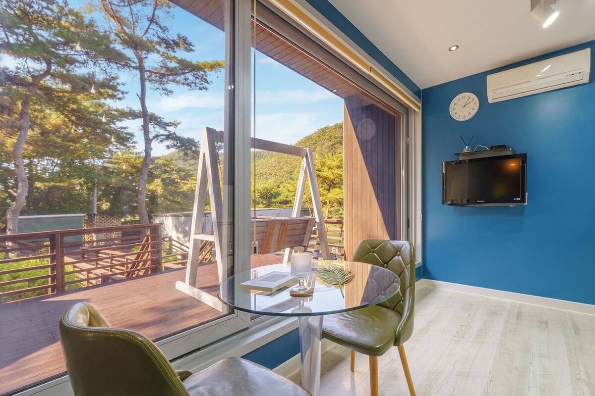 A modern room with a large window, a glass table, two chairs, a television, and an air conditioning unit.