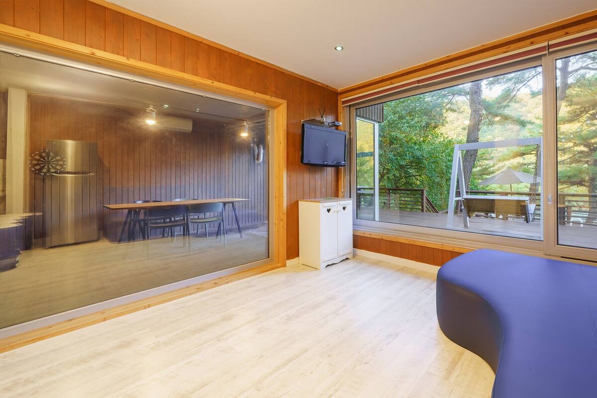 A modern room with wooden walls, a large window with a view of trees, a flat-screen TV, and a dining area with a table and chairs.