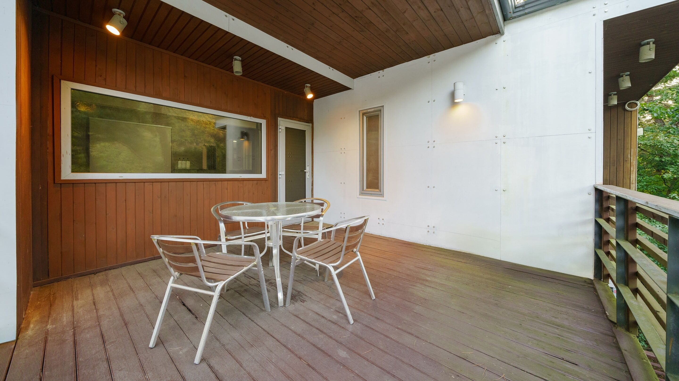 A covered outdoor patio with a wooden deck, a round table with metal chairs, and a view of the surrounding greenery.
