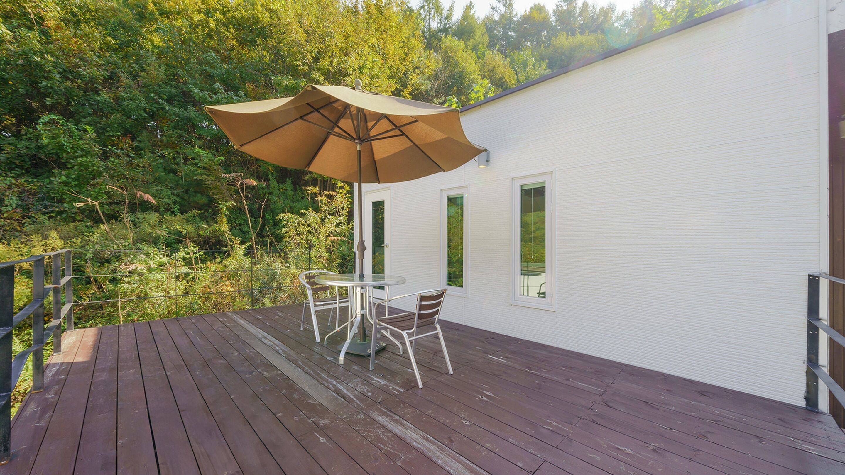 A wooden deck with a table and chairs under a large umbrella, overlooking a forested area.