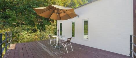 A wooden deck with a table and chairs under a large umbrella, overlooking a forested area.