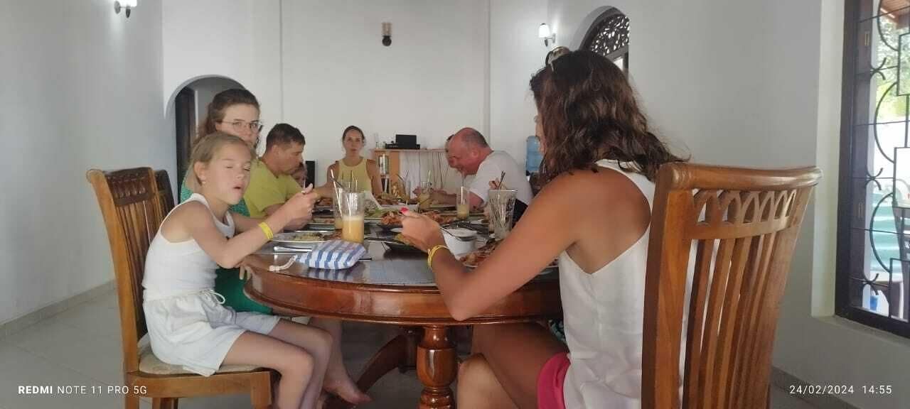 Un grupo de personas sentadas alrededor de una mesa de madera en una habitación con paredes blancas y una ventana con celosía decorativa.