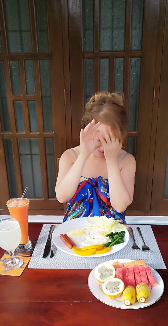 Une personne est assise à une table devant un repas composé d’œufs, de saucisses et de légumes, accompagné d’une boisson et de fruits.