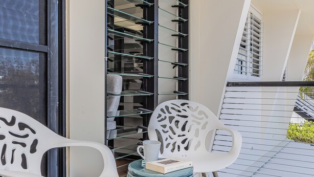 A modern outdoor seating area with white chairs and a green ottoman, a glass shelving unit with books, and a view of a building with balconies. - Beachside Escape by Cottonwood (Casuarina)