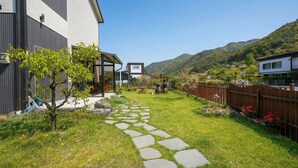 A modern house with a stone pathway leading to a wooden deck, surrounded by lush greenery and a mountainous backdrop.