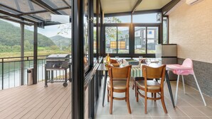 A balcony with a view of a river and mountains, a grill, a table with chairs, and a pink high chair.