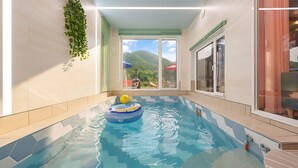 An indoor swimming pool with a blue inflatable float, tiled walls, and a view of a mountain landscape through the windows.