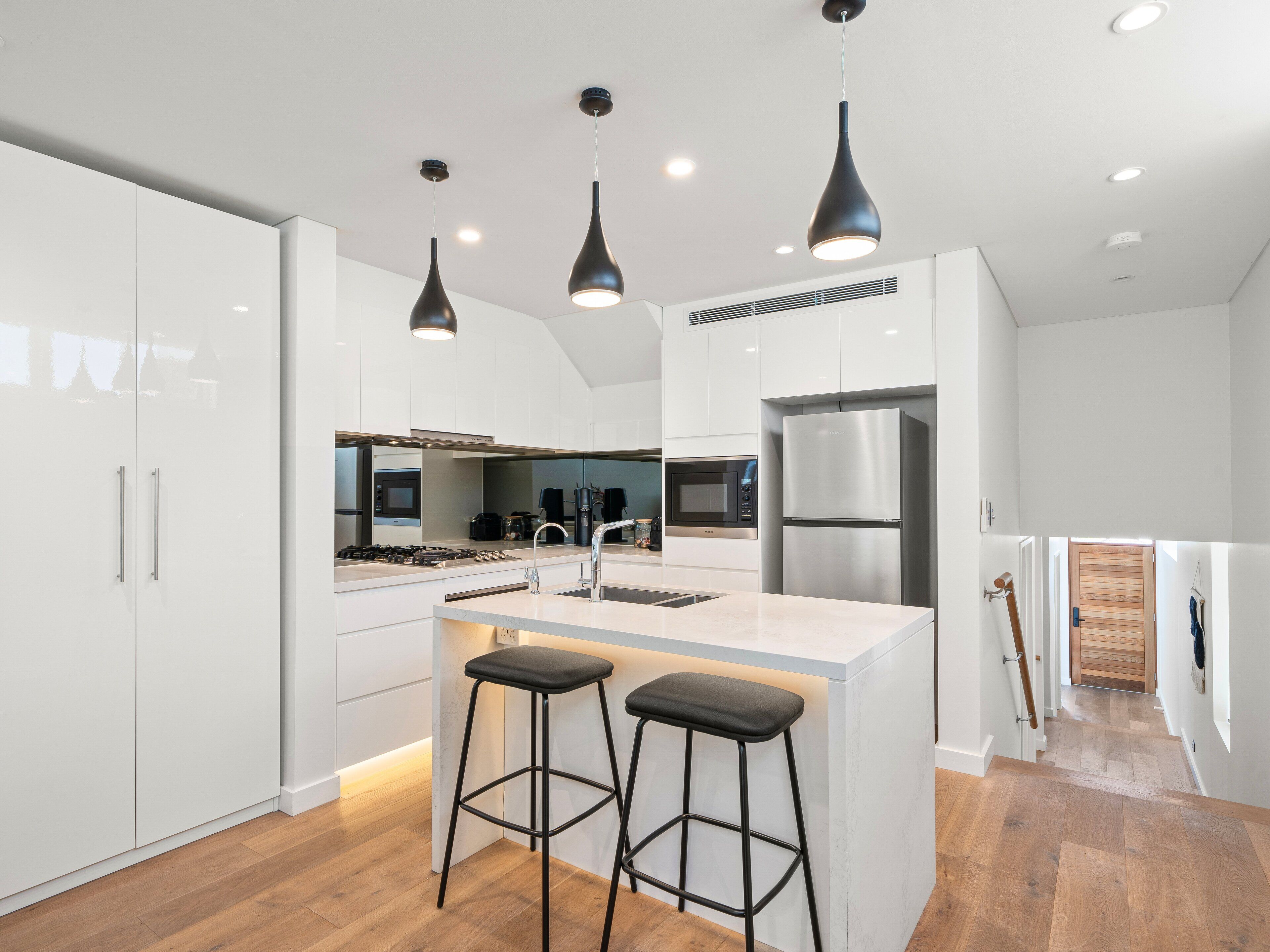 Una cocina moderna con armarios blancos, un refrigerador de acero inoxidable, una isla central con taburetes e iluminación colgante.