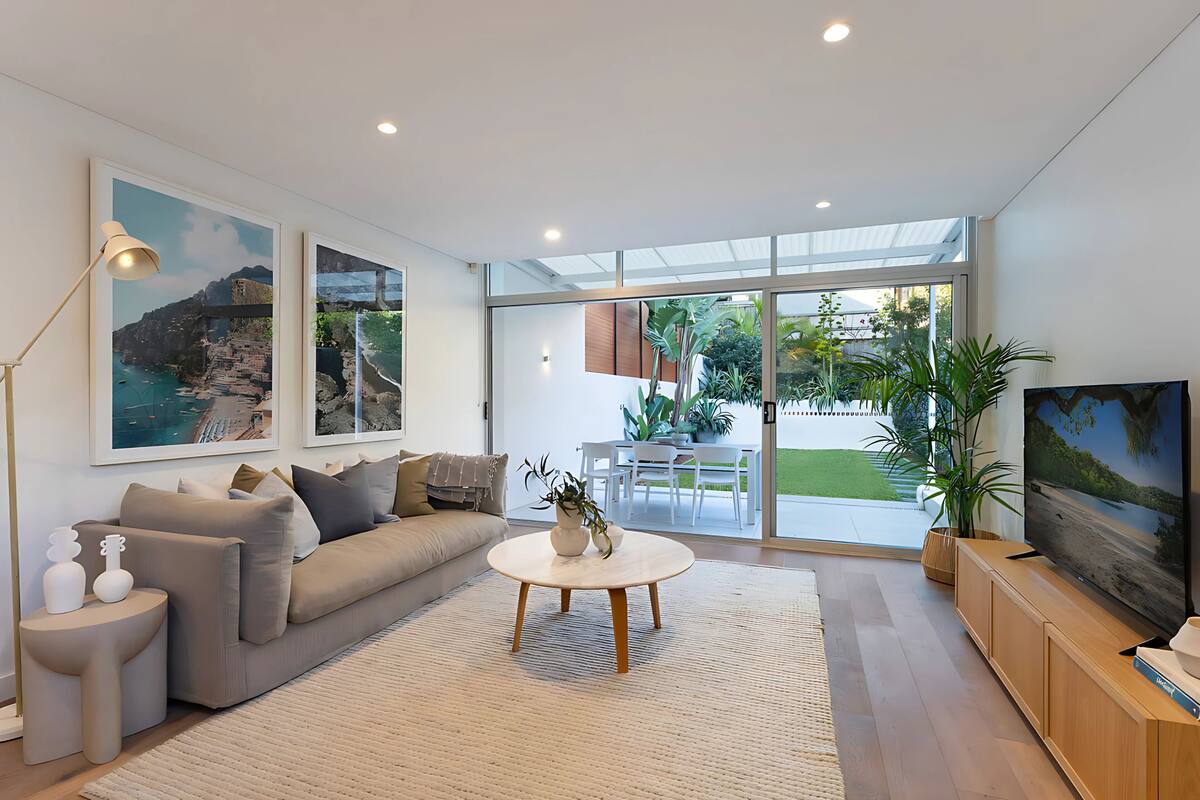 Una sala moderna con un sofá, una mesa de centro y una televisión. Hay cuadros enmarcados en la pared, una planta y vistas al exterior a través de amplias ventanas.