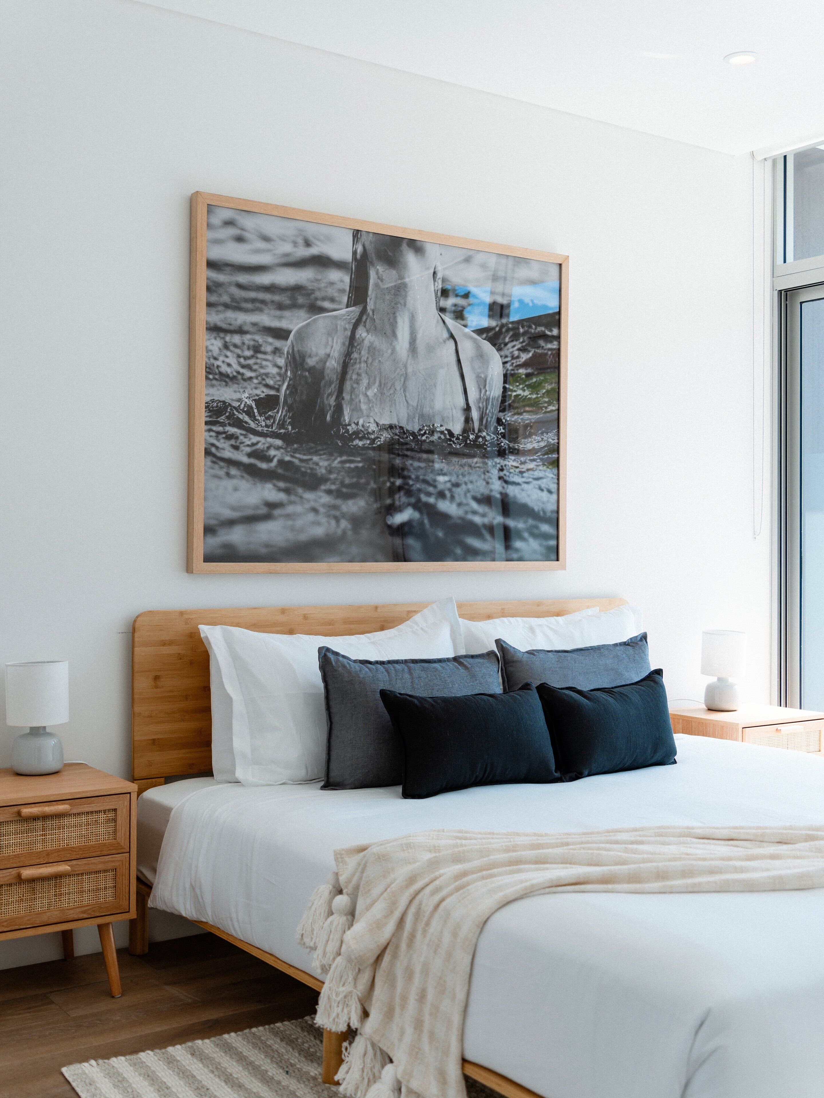 Een slaapkamer met een bed, kussens, een nachtkastje, een lamp en een lijst met een foto aan de muur.