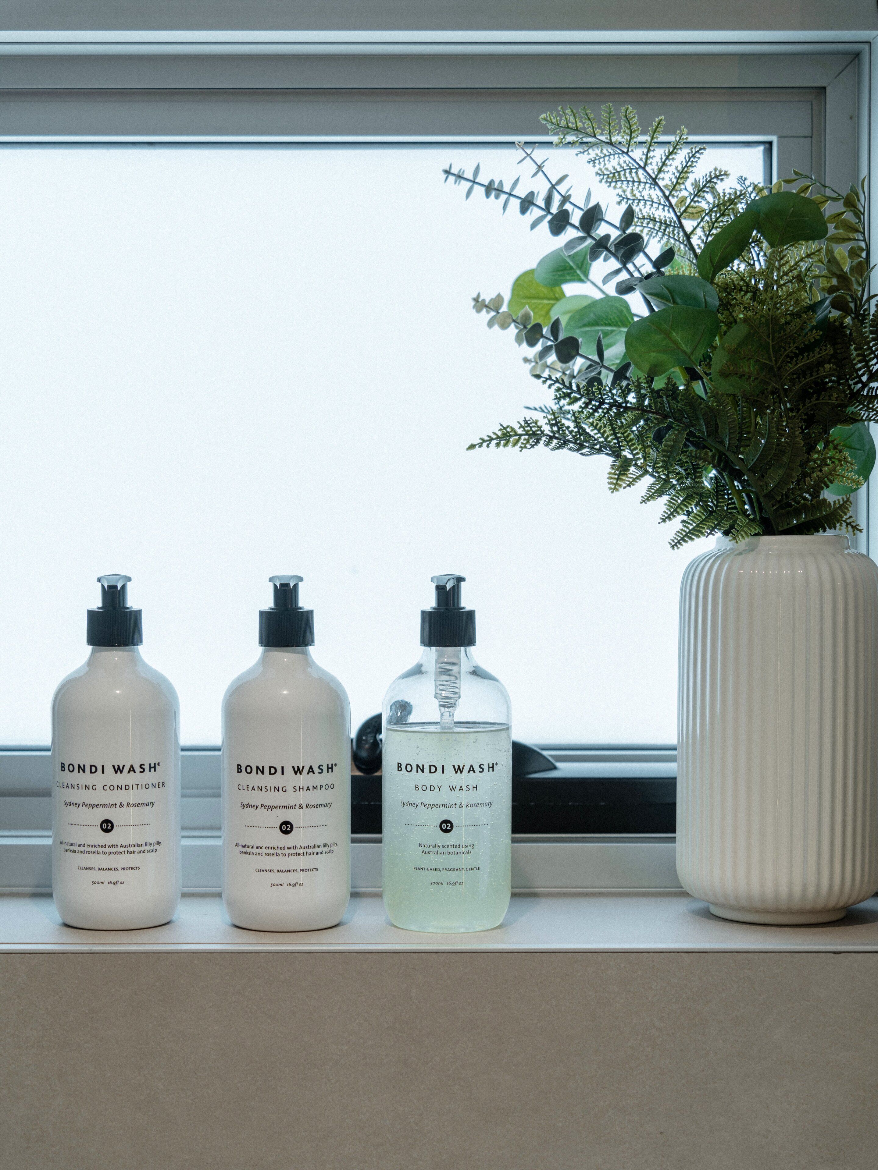 Three bottles of Bondi Wash® on a windowsill, next to a vase with green plants.