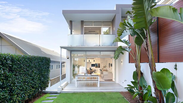 Una casa moderna de dos pisos con fachada de vidrio, una cocina a la vista y un pequeño espacio de estar al aire libre.