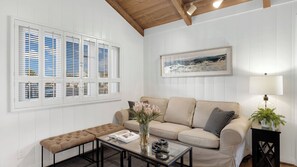 A living room with a beige sofa, a glass coffee table, a bench, a framed picture on the wall, and a lamp on a side table.
