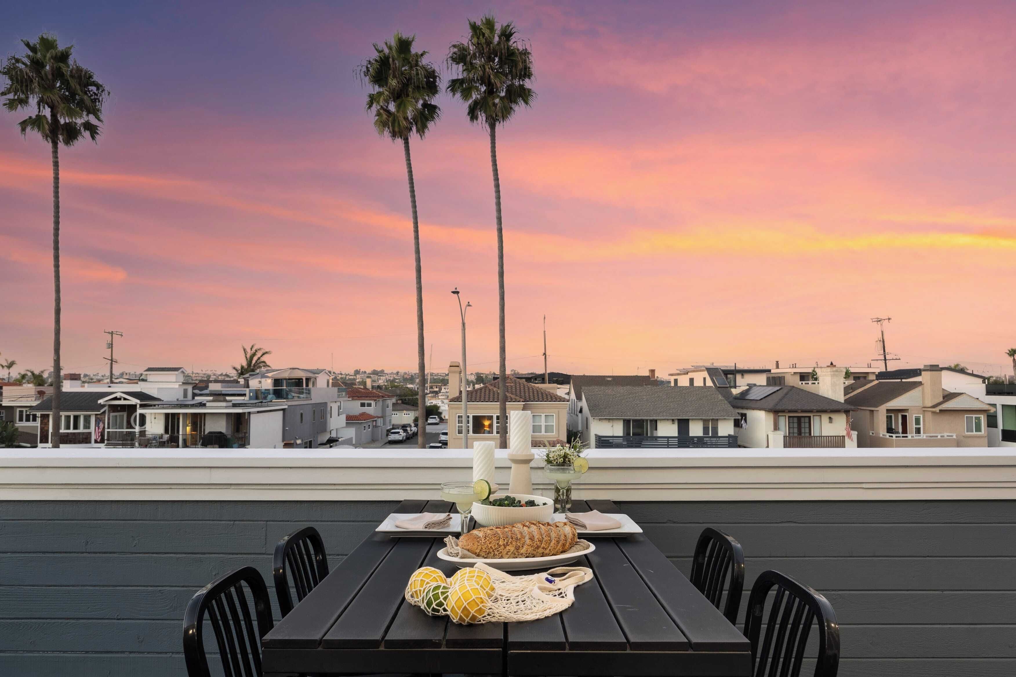 A rooftop dining area with a table set for two, overlooking a residential neighborhood and a sunset sky.