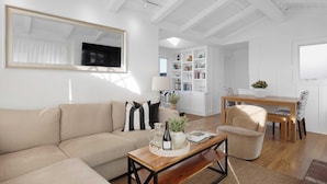 A modern living room with a beige sofa, a wooden coffee table, and a dining area with a wooden table and chairs. - Twin Dolphins by Avantstay Steps to Beach Deck (Newport Beach)