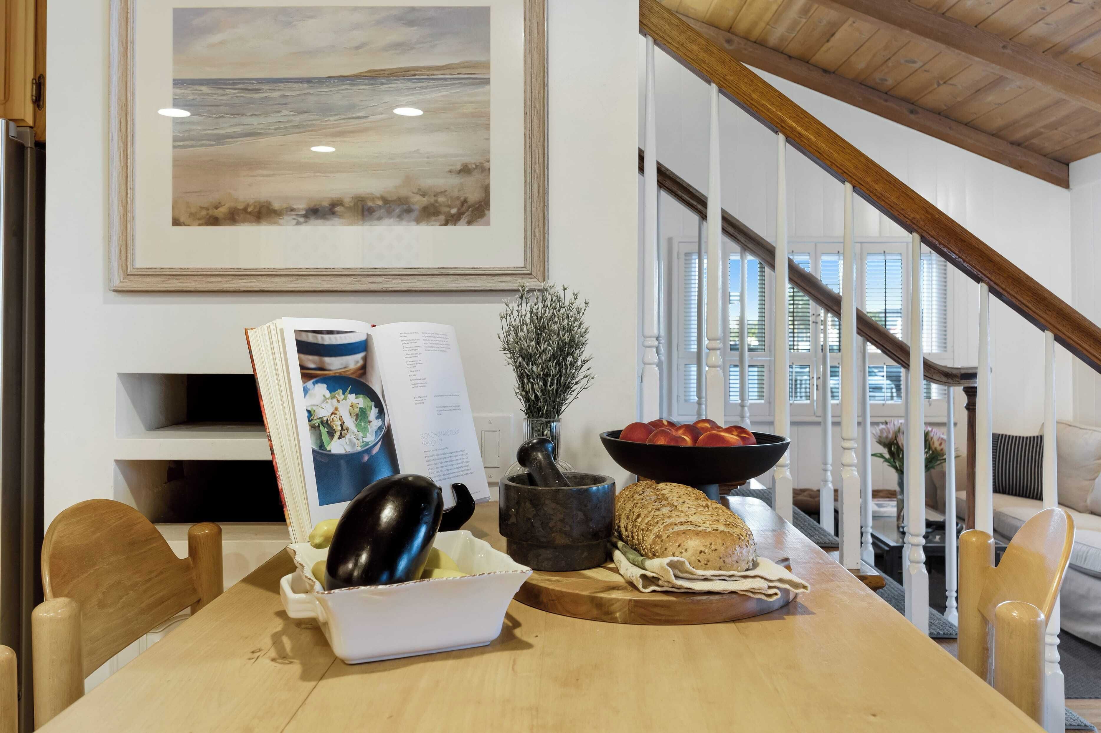 A dining area with a wooden table, a bowl of fruit, a book, and a framed painting on the wall.