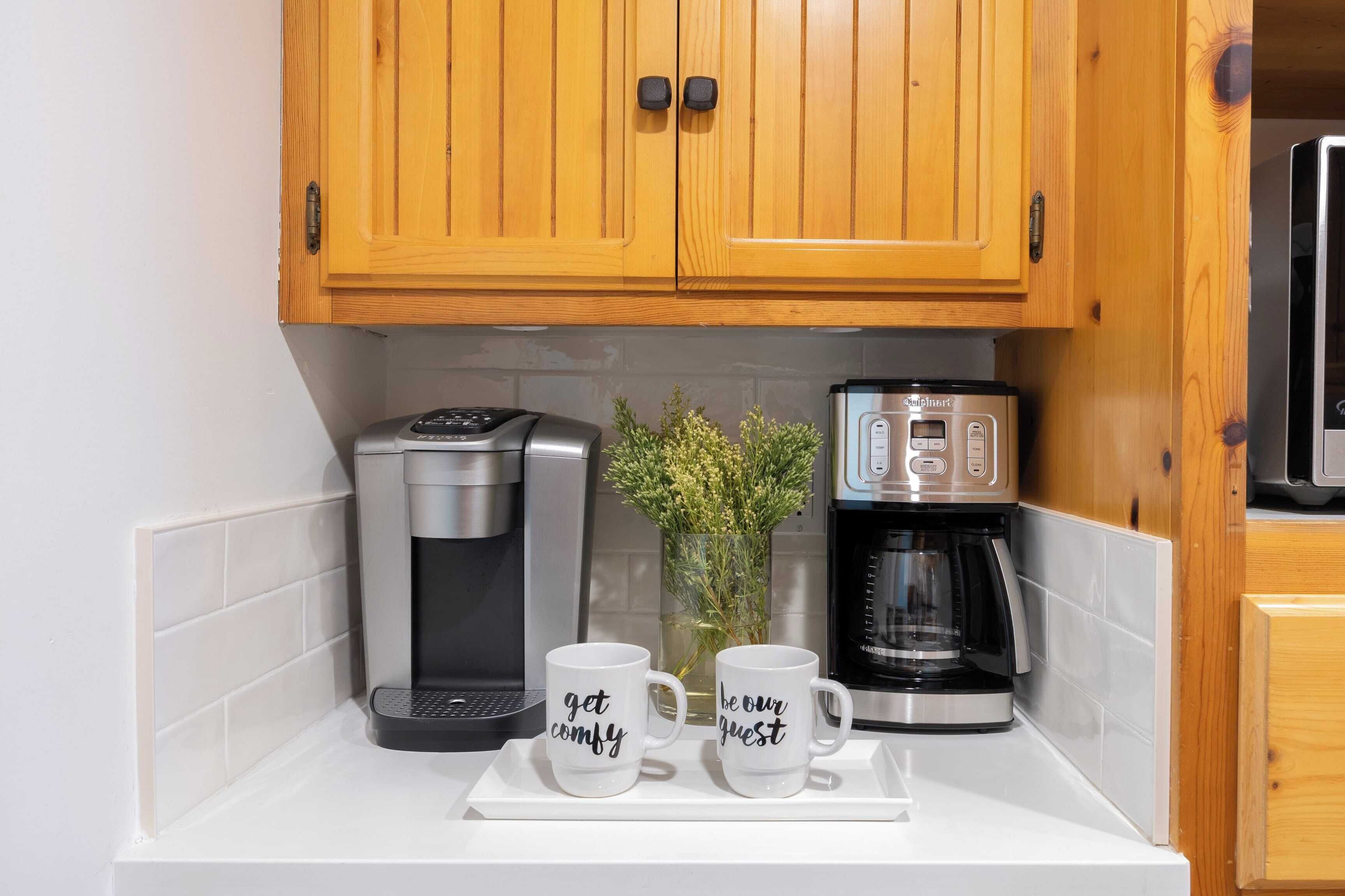 A kitchen with wooden cabinets, a coffee maker, and two mugs with text.