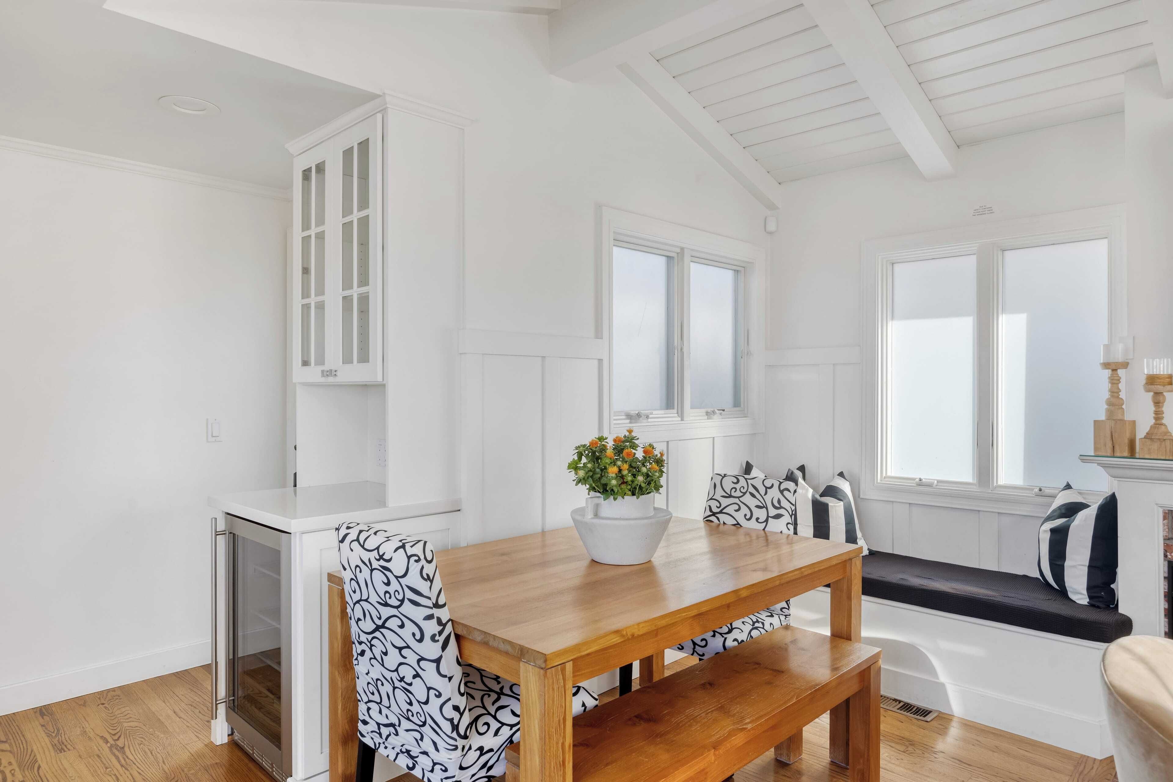 A bright, white interior with a wooden dining table, bench seating, and a potted plant on the table.