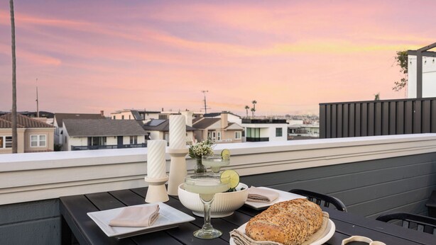A table with a loaf of bread, a glass of drink with a lime, and a vase of flowers on a rooftop terrace with a city view. - Twin Dolphins by Avantstay Steps to Beach Deck (Newport Beach)
