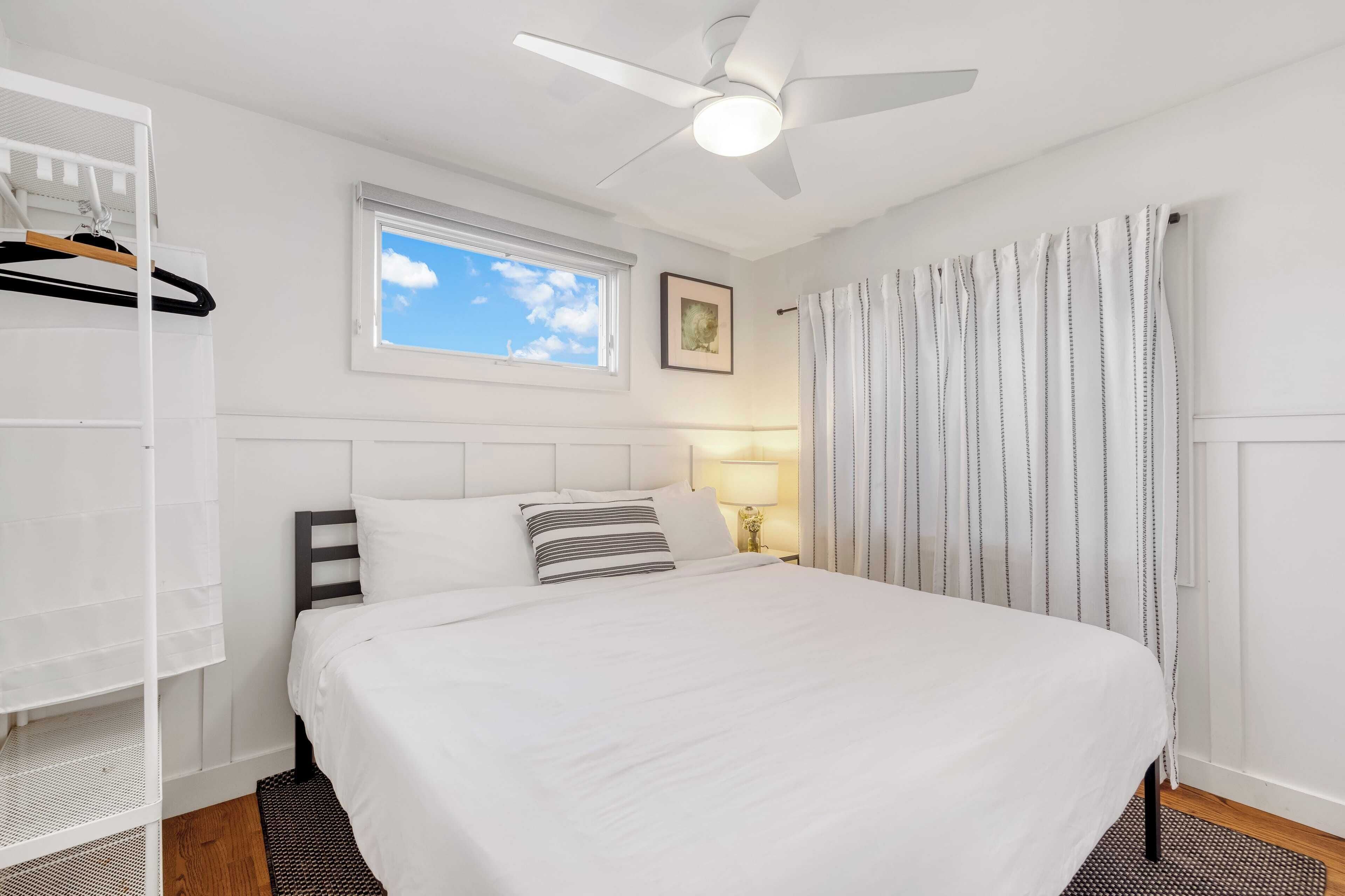 A neatly arranged bedroom with a bed, a ceiling fan, a window with a view, and a bedside lamp.